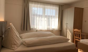 A cozy bedroom with two single beds and a table. Bright curtains let in plenty of light, and the wardrobe offers additional storage space.