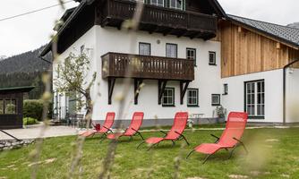 A modern house with a balcony and a wooden facade. In the foreground, there are red loungers on a green meadow.
