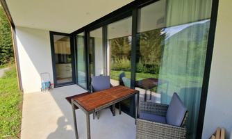A cozy terrace with a table and chairs. The large windows offer a view of the green surroundings.
