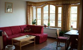 A cozy living room with a red sofa and a wooden table. Large windows bring in plenty of daylight, and fresh plants give the room an inviting atmosphere.