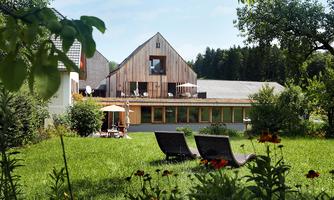 A modern architecture with a wooden building in the background. The garden is green and equipped with deck chairs and a seating area.