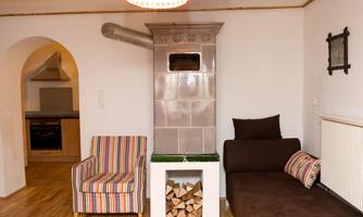 A cozy living room with a tiled stove, a brown sofa, and a striped armchair. The room has light wooden flooring and an open connection to the kitchen.