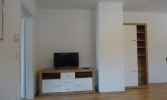 A bright room with wooden flooring and a TV corner. In the background, there is a shelf and a white wall.