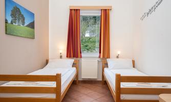 A simple room with two single beds and a window that offers a view of greenery. On the wall hangs a picture of a landscape, and the windows have red curtains.