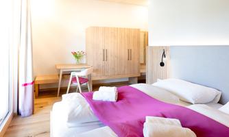 A modern bedroom with a large bed and a purple blanket. The room is bright and cheerful, furnished with a wooden table and a vase with fresh flowers.