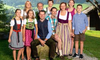 A cheerful group of ten people is wearing traditional costumes. They are standing outdoors in front of a beautiful mountain landscape.