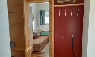 A cozy entrance area with wood paneling and a red wall. In the background, a bright room with a couch and curtains is visible.