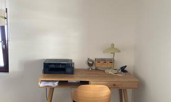 A simple workspace with a wooden desk and a chair. On the table, there is a printer, a lamp, and some office supplies.