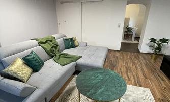 A modern living room with a bright sofa and colorful cushions. A round, green table stands on a patterned rug.