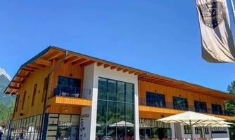 A modern building with wooden cladding and large windows. In front of the building, there are tables and umbrellas.