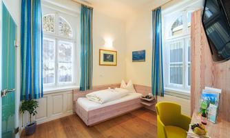 A bright room with large windows and blue curtains. There is a single bed, a desk, and cozy decoration.