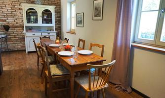 A cozy dining area with a long wooden table and chairs. The walls are light, and there is a charming brick wall as well as windows that let in plenty of light.
