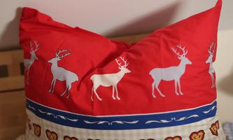 A red pillow with white reindeer motifs and a blue stripe. The upper part of the pillow has a pointed shape.