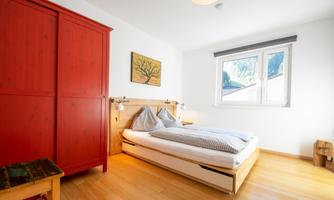 A cozy bedroom with a large bed and a red wardrobe. The room is bright and welcoming with a window that looks out onto nature.