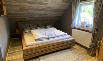 A cozy bedroom with a large wooden bed and a warm wooden ceiling. The window lets in a lot of light, and the room is simply decorated.