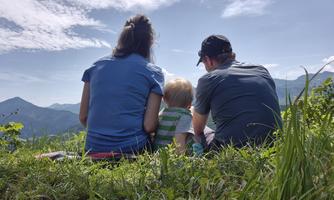 A family is sitting on a meadow and looking at the mountains. The sky is clear and blue.