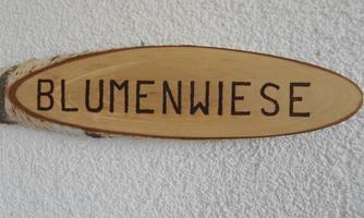 A wooden sign with the inscription "FLOWER MEADOW". The sign has an oval shape and is light brown.