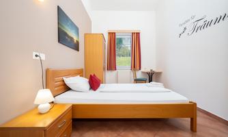 A cozy room with a single bed, wooden desk, and a small table. A picture hangs on the wall and there are bright curtains that let in a lot of light.
