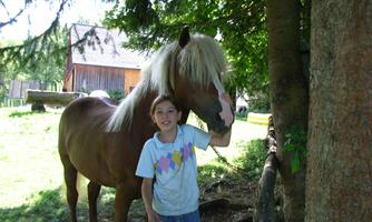 A boy stands next to a horse under trees. In the background, a rural house is visible.
