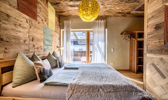 A cozy bedroom with wooden walls and a large window. There is a big bed with pillows and a striking yellow lamp.