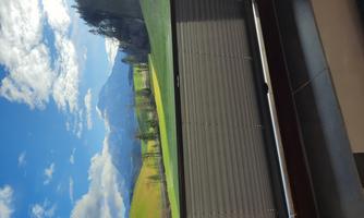 A window view of a green landscape with mountains in the background. The sky is blue with some clouds.