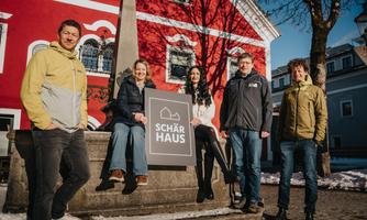 A group of five people stands in front of a building with a red facade. They are holding a sign that says "SCHÄR HAUS".