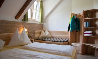 A cozy room with two beds and natural wooden elements. Bright curtains let light in and a small seating area invites you to relax.