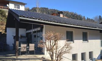 A modern, white house with a black roof in a mountainous area. There is a terrace with garden furniture and a clear, sunny sky.
