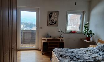 A cozy bedroom with a large bed and a desk. Through the windows, you can see the mountains and a balcony.