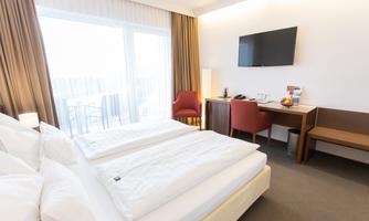 A modern hotel room with a double bed and a cozy seating area. The large window area lets in plenty of light and provides access to a balcony.