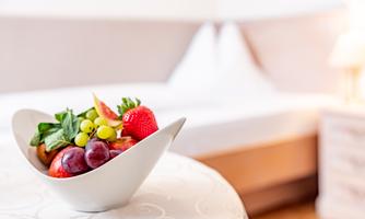 A bowl of fresh fruit is on a table. In the background, a bed and a cozy atmosphere can be seen.