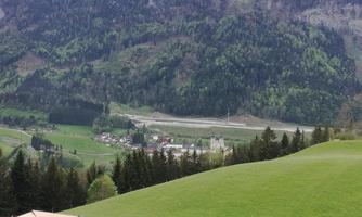 A picturesque landscape with green meadows and mountains in the background. In the valley lies a small village, surrounded by trees.