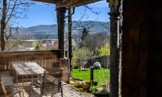 A cozy wooden terrace with a table and chairs that offers a view of the green landscape. In the background, mountains and a blue sky can be seen.