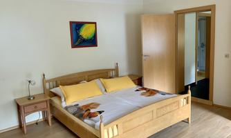 A bright bedroom with a double bed and a bedside table. A colorful picture hangs on the wall, and an open door leads to a hallway.