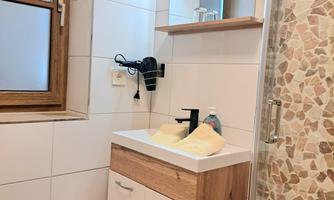 A modern bathroom with a wooden sink and white tiles. Light pours in through the window and the shower cabin is visible.
