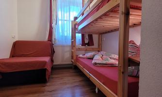 A cozy room with a wooden bunk bed and a sofa bed. The room has windows with curtains and a warm wooden floor.