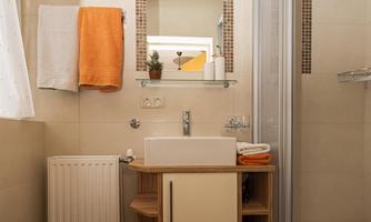 A modern bathroom with a shower, sink, and wooden shelf. Towels in orange provide a colorful accent.