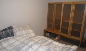 A simple bedroom with a bed and a wooden display cabinet. The bedding has a checked pattern, and the room appears tidy and minimalist.