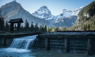 A beautiful waterfall flows over a wooden structure, surrounded by snow-covered mountains and a forest. The clear sky makes the landscape shine.
