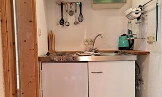 A small kitchenette with a sink, a stovetop, and a refrigerator. Above the countertop are shelves and kitchen utensils.