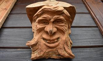 A wooden sculpture of a smiling man with a beard and hat. The figure is attached to a wooden wall.