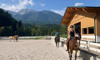 A riding arena in nature with mountains in the background. Two female riders are riding on their horses, while another horse is standing in the background.