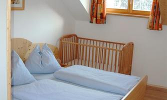 A bright bedroom with two single beds and a crib. The room features wooden details and large windows with colorful curtains.