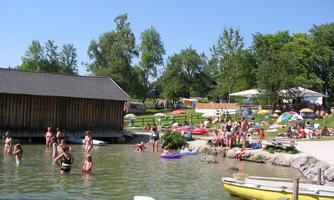 A lively bathing lake with many people swimming and sunbathing. Around the lake, there are boats and loungers on a green meadow.