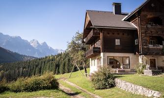 A cozy wooden house amidst a picturesque mountain landscape. In the background, majestic mountains and a clear blue sky are visible.