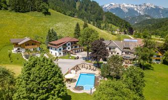 A picturesque landscape with several buildings, surrounded by green meadows and trees. In the foreground, there is a swimming pool and in the background, mountains can be seen.