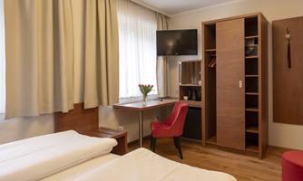 A cozy room with two single beds, a desk, and a television. The room has large windows with curtains and a wardrobe for storage.