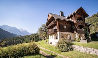 A cozy wooden house in a green landscape, surrounded by mountains. The clear sky makes nature shine.