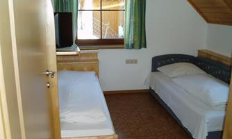 A simple bedroom with two beds and a window with green curtains. The room has a wooden floor and a bright, cozy atmosphere.