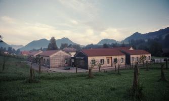 A rural scene with modern buildings and gentle hills in the background. The sky is cloudy, and the surroundings are calm and green.